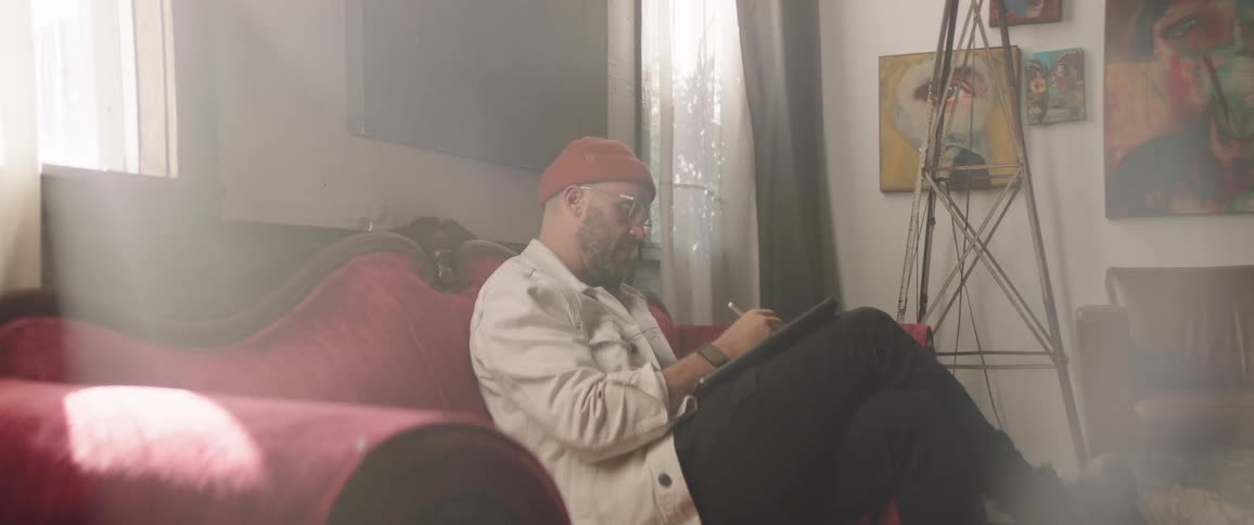 Artist sits on couch as sun rays shine through windows drawing on tablet