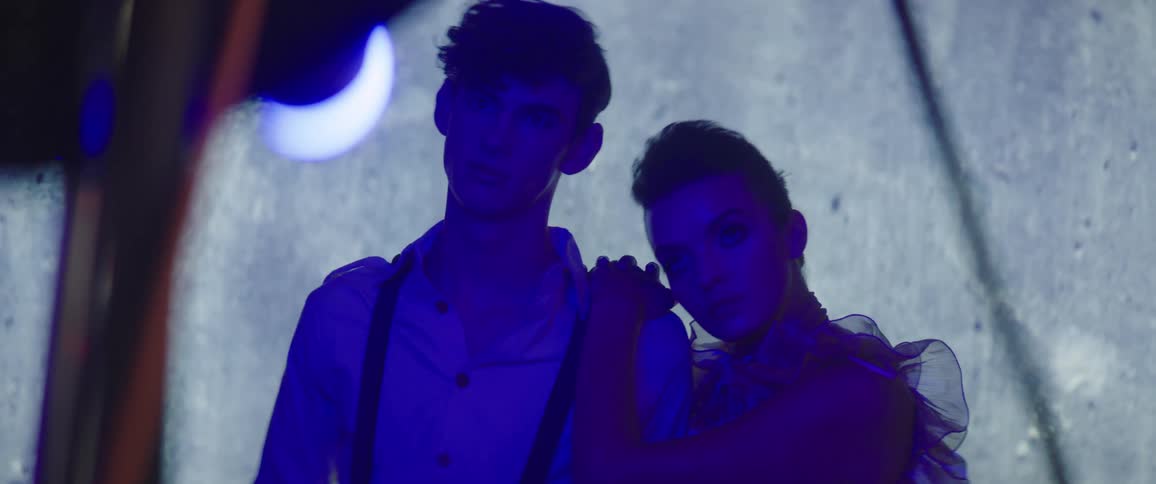 Fashion model pose together the woman leaning on the man under colored lighting as the camera flashes