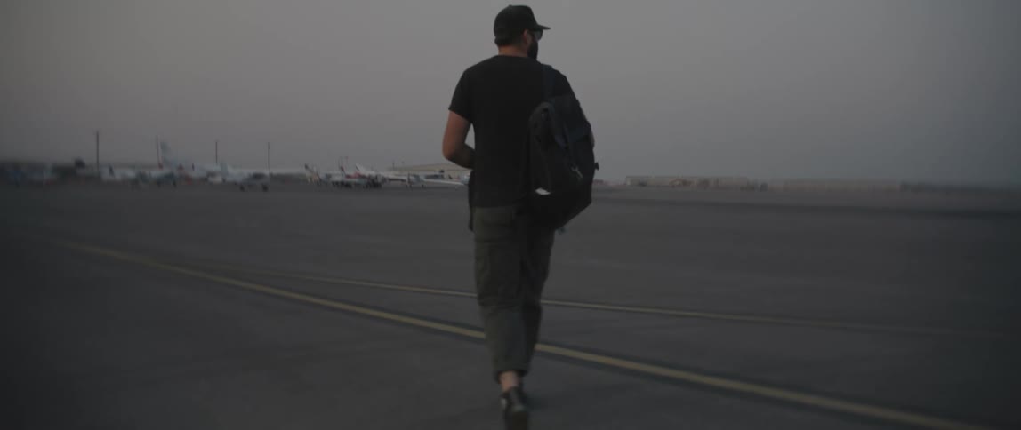 A man with a hat and a backpack walking on a runway with airplanes the sky and clouds in the background