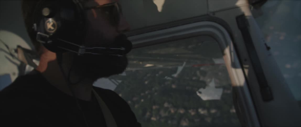 Bearded man wearing sunglasses piloting an airplane over a small town at evening