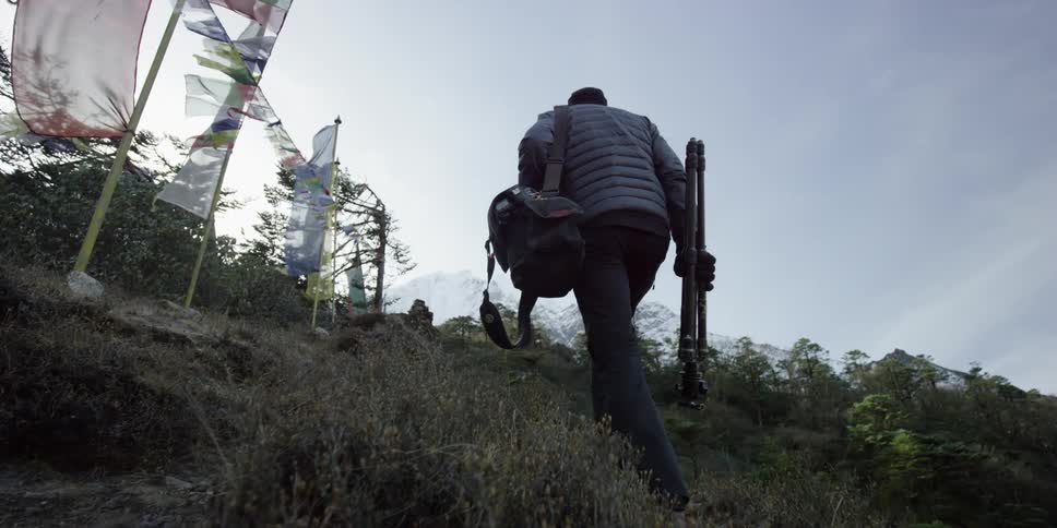 A man with camera bag and tripod climbs a hill toward some banners