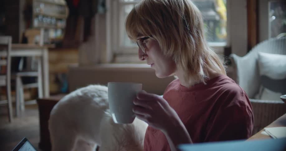 Blonde woman with glasses and red t shirt drinks a cup of coffee and pets golden retriever puppy in the living room at home