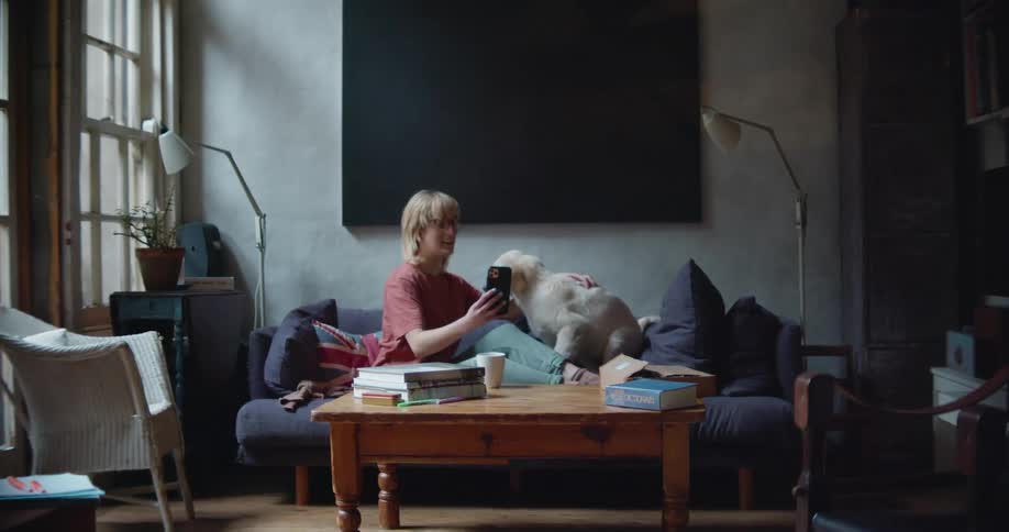 A woman sitting with a puppy on a sofa in a living room at home taking a selfie with a smart phone