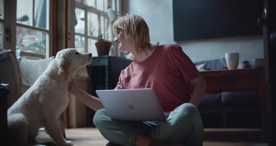 Blonde woman sitting cross legged on floor using laptop briefly pets dog