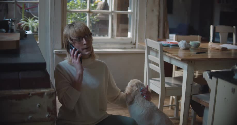A woman with glasses in a room talking on a smart phone and petting a dog
