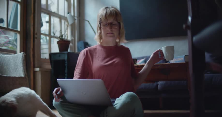 A woman sitting on the floor in a living room with an apple laptop computer in her lap next to a light brown dog taking a sip of coffee from a mug