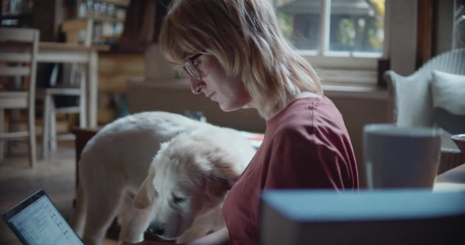A woman with glasses sitting on the floor using a laptop and petting a puppy
