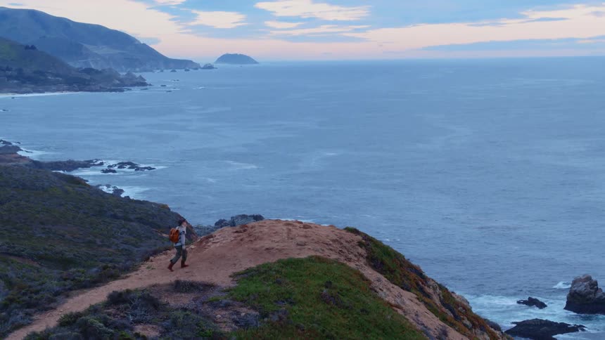 A woman is hiking toward the edge of a cliff to view the ocean at dusk
