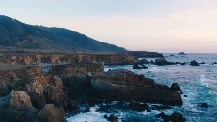 Waves are crashing at the base of cliffs at dusk