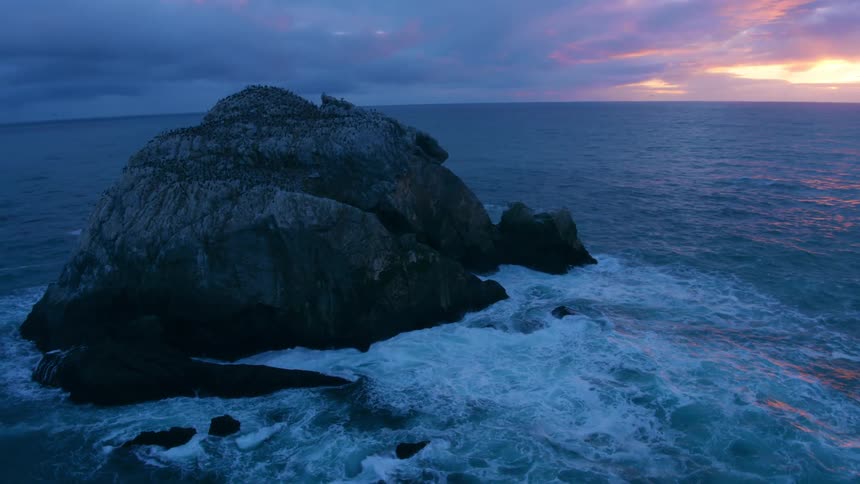 It is a rock in the ocean and the skies are stormy and the water is crashing against the rock