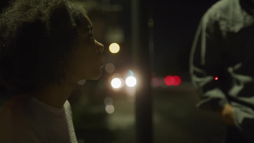 A woman sits outside on a bench an nervously looks up as a man approaches