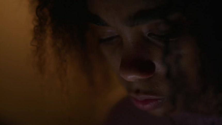 A woman closes her eyes and looks down as she stands in a dim room