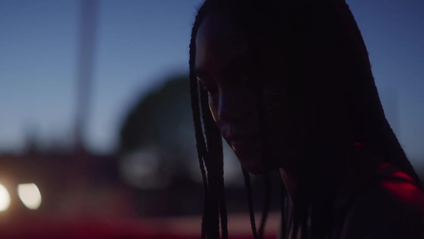 The silhouette of a young woman with braids can be seen as she stands outside at night