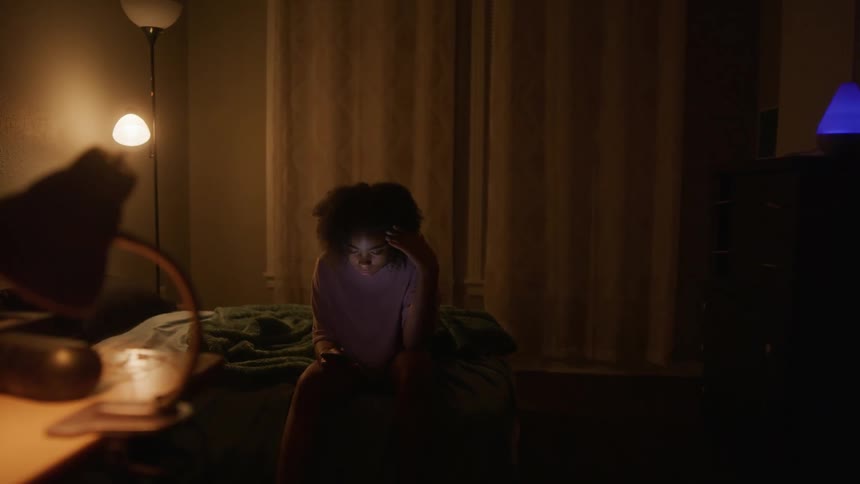 Young black woman sits on bed, looking at cell phone