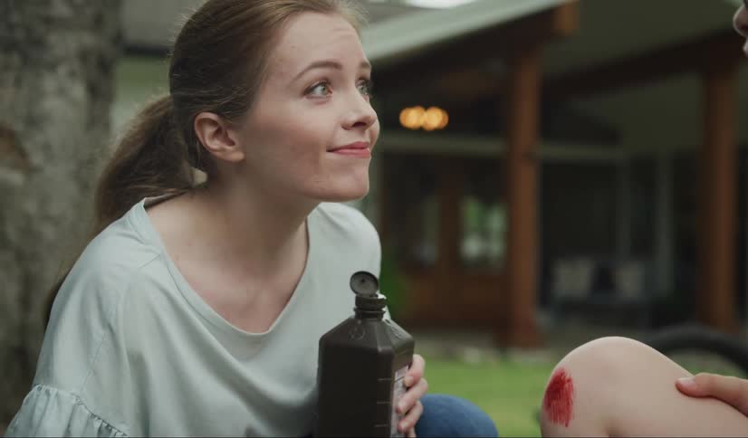 A woman putting hydrogen peroxide on a boys cut on his knee with a house the sky and clouds in the background