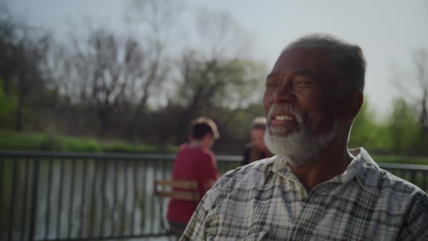A man laughs after moving a chess piece while playing with a friend on a park patio over a lake