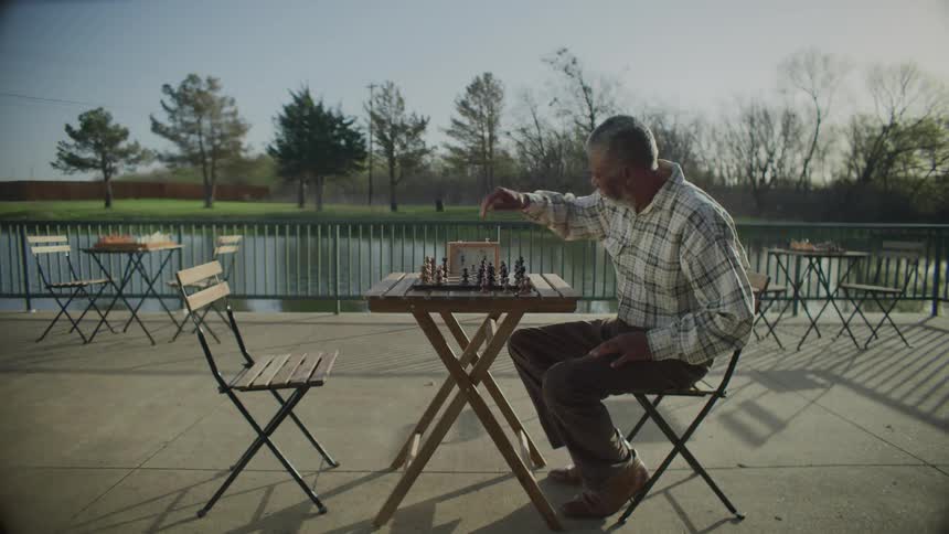 A man sitting on a chair by a table playing chess with a pond trees the sky and clouds in the background