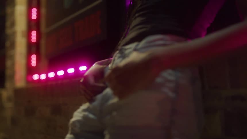 Man leans against a store front at night with his thumbs hooked into the belt loops of his jeans