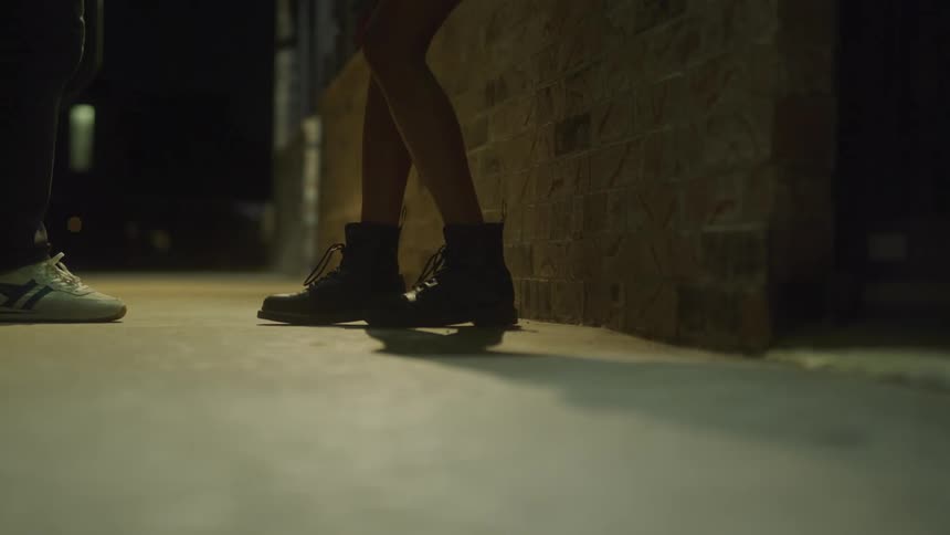 Woman wearing shorts waits by brick wall at night and is joined by man in jeans before walking off with him