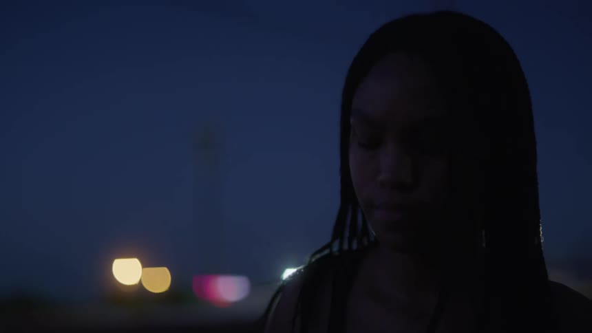A young woman with braids stands outside at night near to a building with bright lights