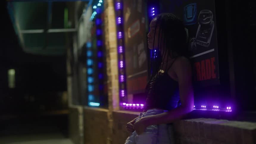 A young woman leans against the front of a building with colorful flashing lights around its windows
