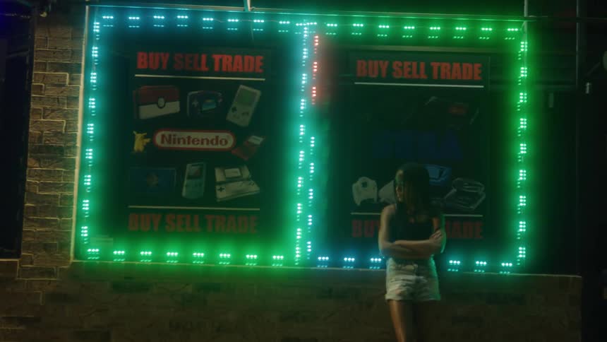 A woman stands with her arms crossed in front of a neon side on the side of a brick building