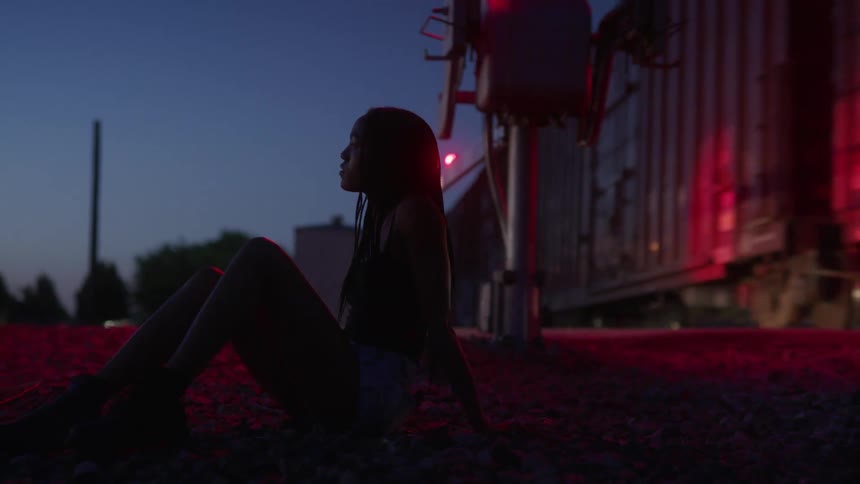 A young woman glances around as she sits on the ground near train tracks as a freight train passes by just a few feet behind her