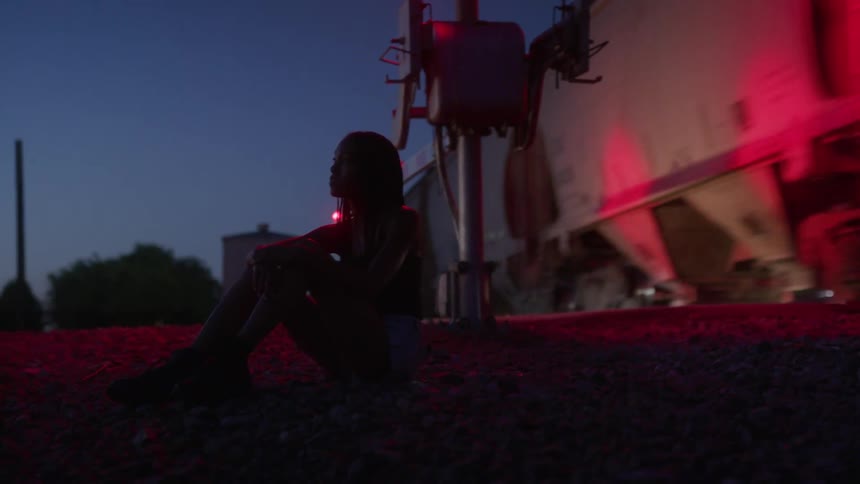 A young woman sits on the ground next to train tracks as a freight train passes by at night