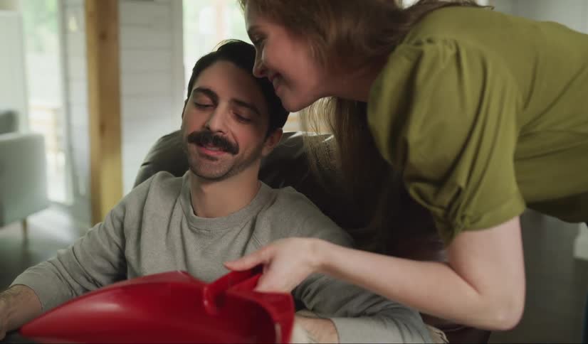 A man sitting on a couch as a woman walks up and kisses him and hands him a dustpan