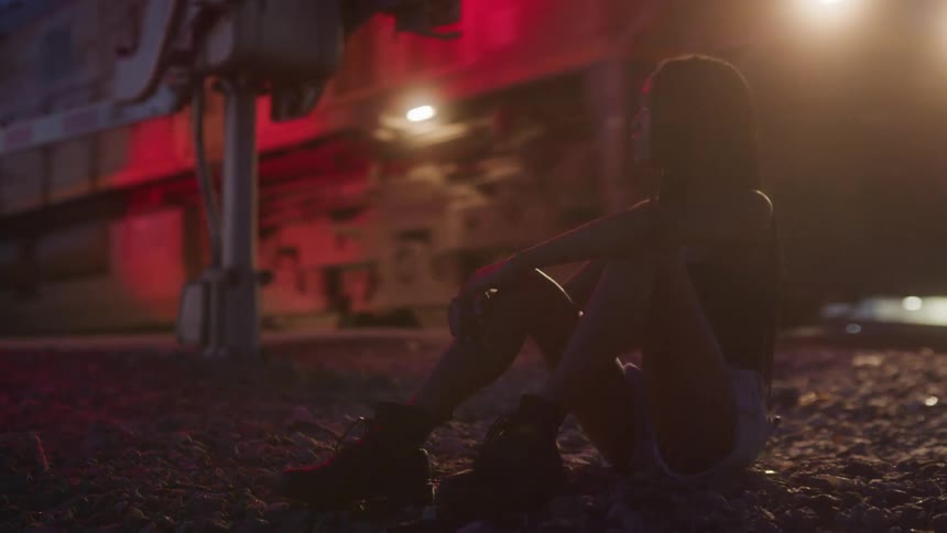 A girl sits near the train crossing barrier and turns her head to watch the train go by