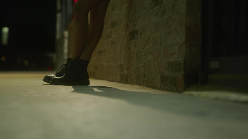 A female wearing boots leans against the brick wall of a building