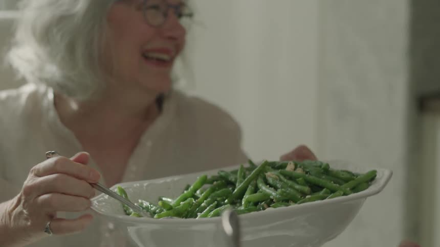 Elderly woman uses a spoon to serve herself some green beans from a bowl then she passes the bowl to another woman