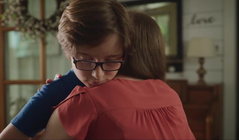 A depressed boy adjusts his glasses before he is hugged by a woman and begins to smile