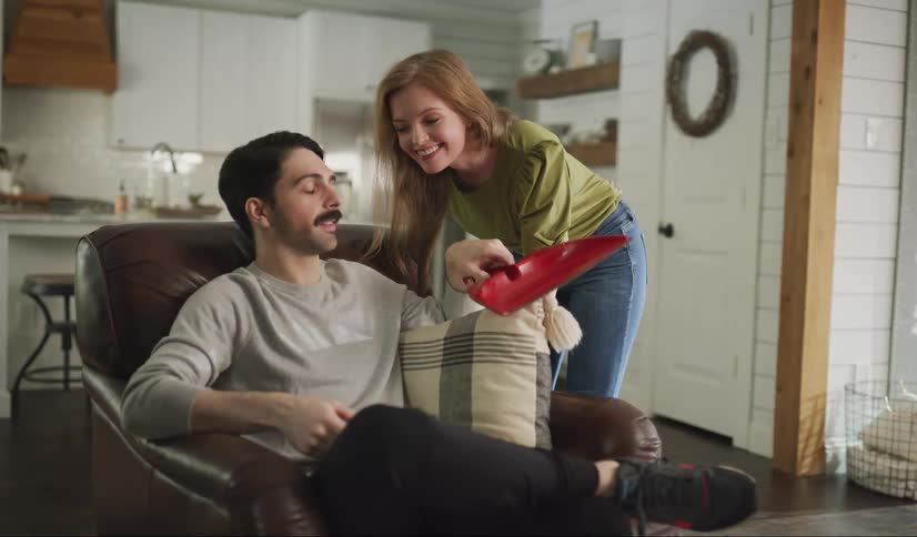 A young woman walks over and kisses her significant other then hands him a dustpan to use to clean up the kitchen