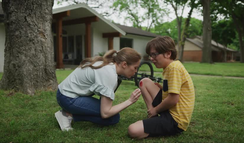 A woman putting hydrogen peroxide on a boys cut on his knee with a house trees the sky and clouds in the background