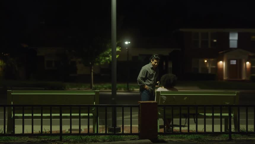 Man walks along sidewalk carrying mobile phone stopping to speak to woman sitting on bench across street from house
