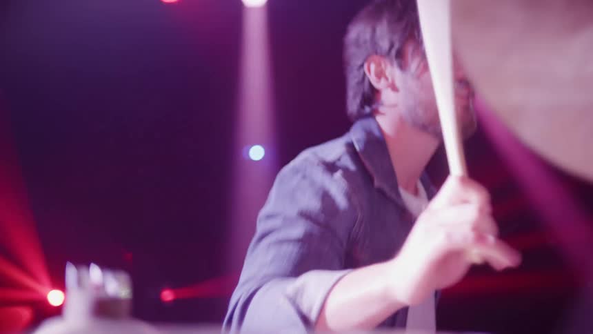 A man in a stage playing drums with drumsticks with lights in the background