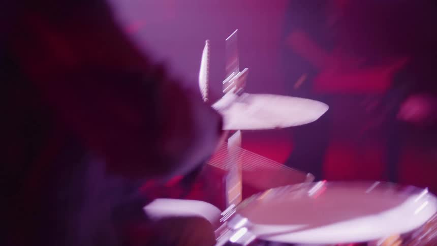 A man on stage playing drums with drumsticks with a woman playing a keyboard in the background