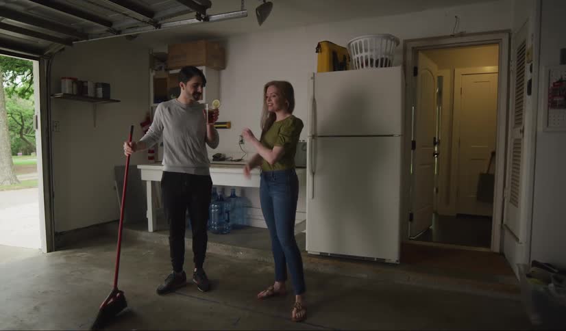 Man with a moustache sweeps the garage floor and woman bring him drink appreciates him