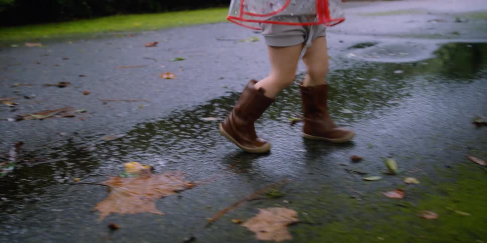 A little girl wearing shorts and rainboots walks on a wet driveway