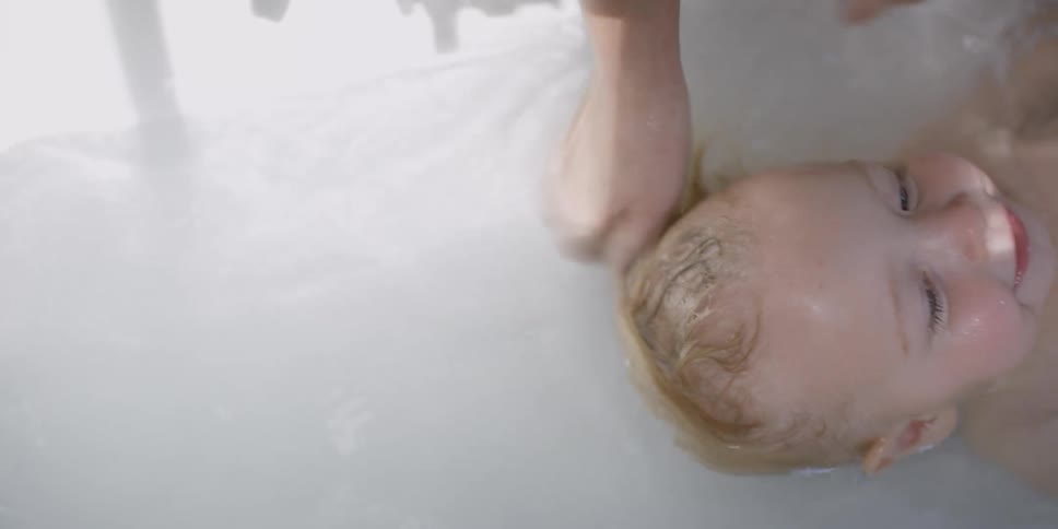 A person in a bathroom bathing a little girl in a bathtub with water
