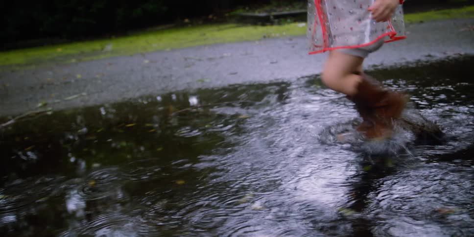 A little girl with boots running in a puddle of water with trees the sky and clouds in the background