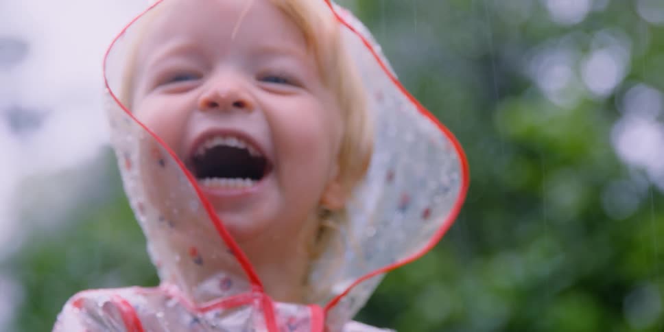 Blonde girl wearing clear rain coat decorated with flowers laughs standing outside as it rains