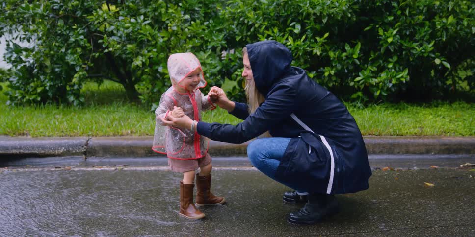 A kneeling woman holds the hands of a young girl as the girl jumps in the water running down a road on a rainy day