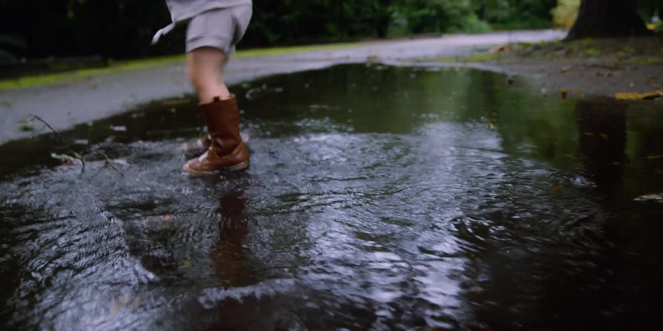 A child with boots stepping on a puddle of water with trees and plants in the background