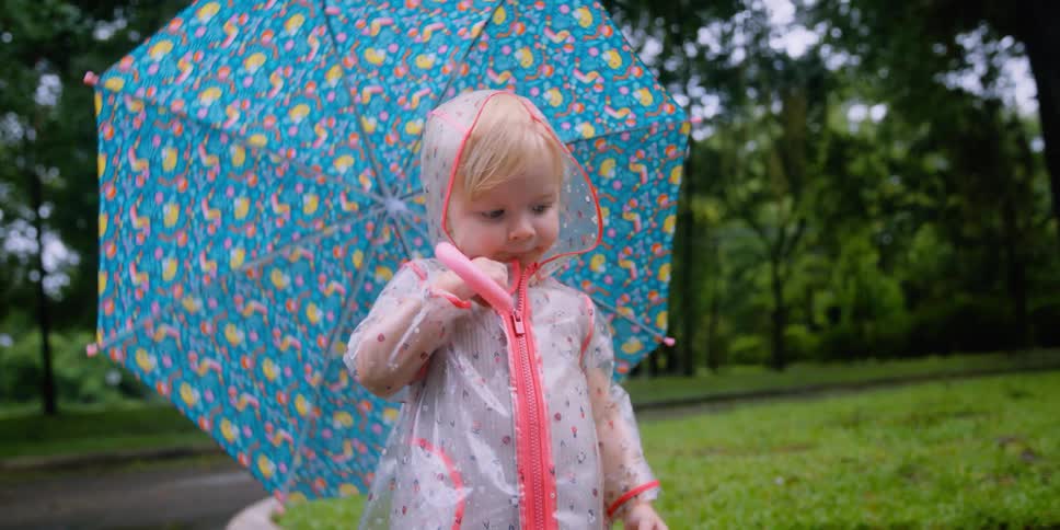 Mother follows daughter wearing raincoat and carrying umbrella as daughter splashes in reflective puddles