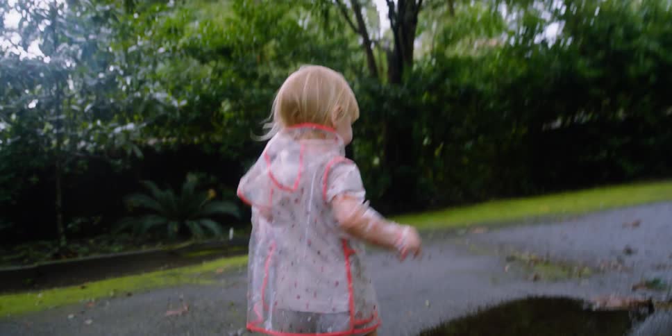Toddler blonde girl with a transparent raincoat and brown boots jumps through a puddle in the park