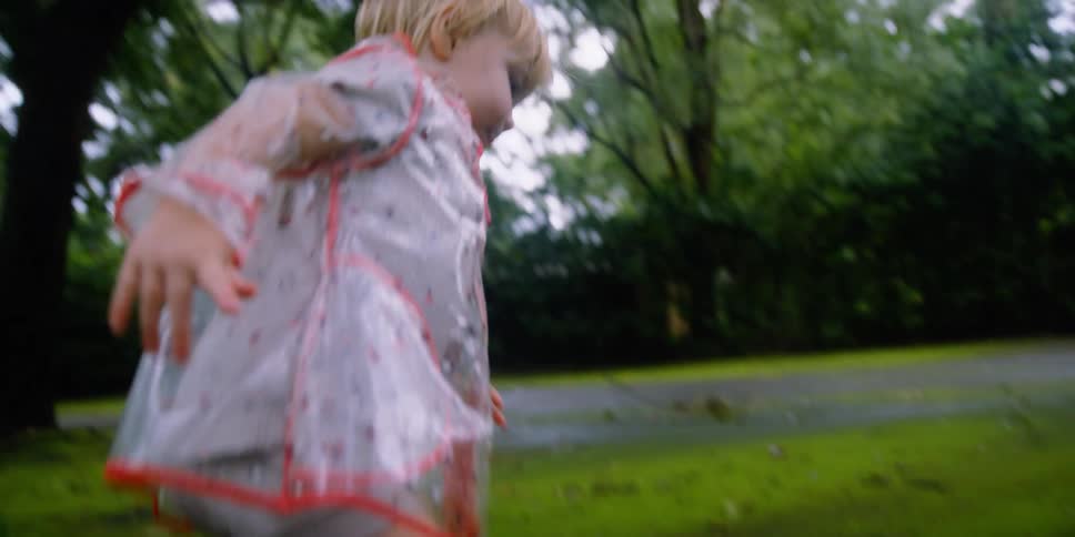 A little girl with boots and a raincoat standing on a concrete slab with trees the sky and clouds in the background