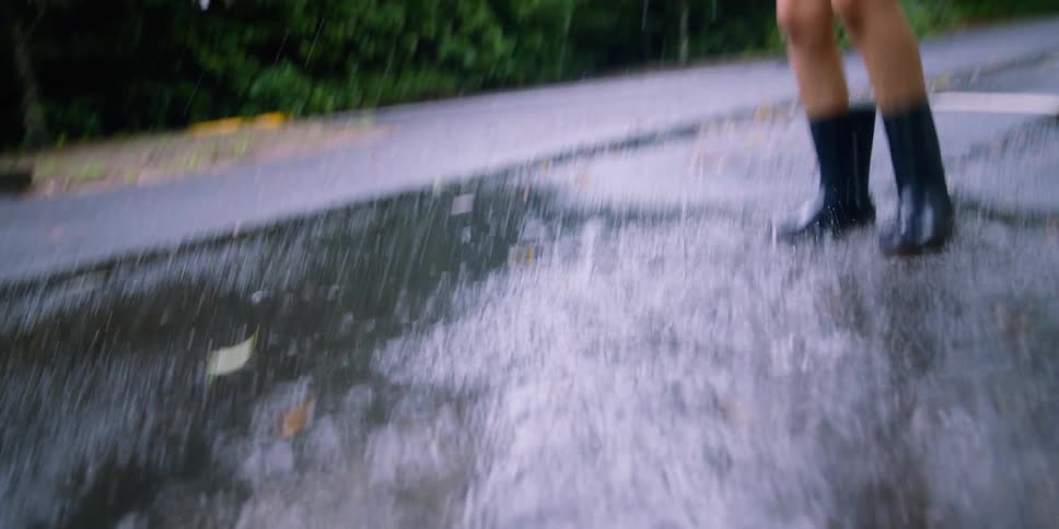 Young boy wearing rubber boots runs and jumps into a puddle of water