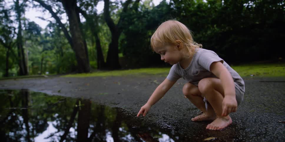 Blonde girl crouches to dip finger in reflective puddle